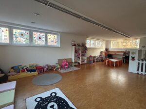 Herna v Dětské skupině Dubina – pohovky s polštáři, herní kobereček, regály s hračkami a dětské stoly pod okny s výzdobou