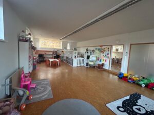 Celkový pohled na hernu Dubina – hrací koutek, regály, hračky, koberec s medvědem a výtvarná stěna s dětskými obrázky